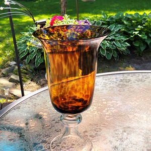 May include: A glass hurricane candle holder featuring a wide, flared rim and a bowl with a mottled, amber and dark brown tortoiseshell pattern. The piece is supported by a clear, transparent glass pedestal base with a rounded, thick stem.
