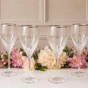 May include: A set of four clear glass wine glasses featuring vertical cut patterns on the bowls and a thin silver rim at the top. Each glass has a slender stem and a circular base, designed for serving beverages with a refined and elegant appearance.
