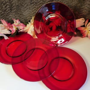 May include: A set of red glass plates, comprising a large serving plate and several smaller plates. The plates are arranged on a white surface, with pink and white flowers in the background. The plates have a glossy finish.