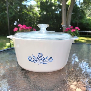 Op de afbeelding: Witte keramische ovenschaal met een blauw bloemenmotief en een glazen deksel. De schaal staat op een glazen tafel.