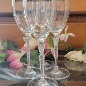 Puede incluir: Un juego de cuatro copas de vino transparentes con bordes plateados. Los tallos tienen un diseño retorcido y las copas están dispuestas sobre una superficie reflectante. Arreglos florales son visibles en el fondo.