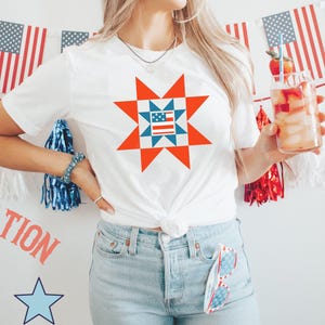 May include: A white t-shirt with a red, white, and blue star-shaped design. The design features a smaller American flag in the center of the star. The shirt is being worn by a person with blonde hair and is paired with blue jeans.