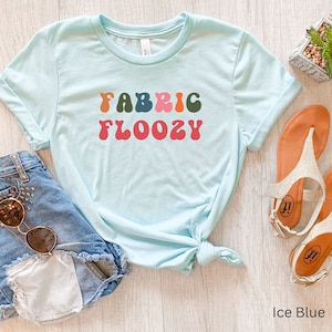 Pu&ograve; includere: Una maglietta azzurro ghiaccio con la scritta "FABRIC FLOOZY" in lettering multicolore in stile retr&ograve;. La maglietta &egrave; abbinata a pantaloncini di jeans e occhiali da sole, e un paio di sandali. La maglietta &egrave; annodata in vita.
