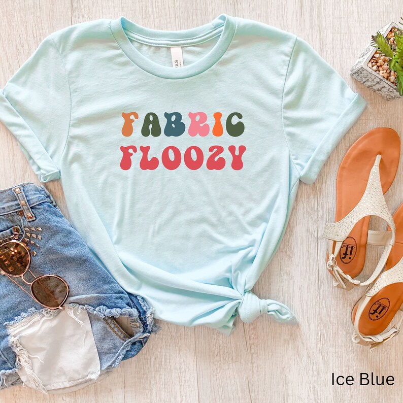 Pu&ograve; includere: Una maglietta azzurro ghiaccio con la scritta "FABRIC FLOOZY" in lettering multicolore in stile retr&ograve;. La maglietta &egrave; abbinata a pantaloncini di jeans e occhiali da sole, e un paio di sandali. La maglietta &egrave; annodata in vita.