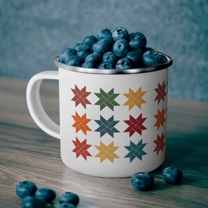 May include: A white enamel mug filled with fresh blueberries. The mug features a colorful star pattern in shades of red, orange, green, yellow, and blue. The mug has a silver rim and a white handle. Several blueberries are scattered on the wooden surface.