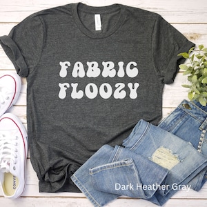 Pu&ograve; includere: T-shirt grigio scuro melange con la scritta bianca "FABRIC FLOOZY", abbinato a jeans blu e scarpe da ginnastica bianche in tela. La maglietta &egrave; realizzata in materiale morbido ed &egrave; adatta per un abbigliamento casual.