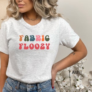 Pu&ograve; includere: T-shirt grigio chiaro con la scritta "FABRIC FLOOZY" in un font retr&ograve; colorato. La maglietta &egrave; a girocollo. La parola "Ash" &egrave; scritta in basso a destra.