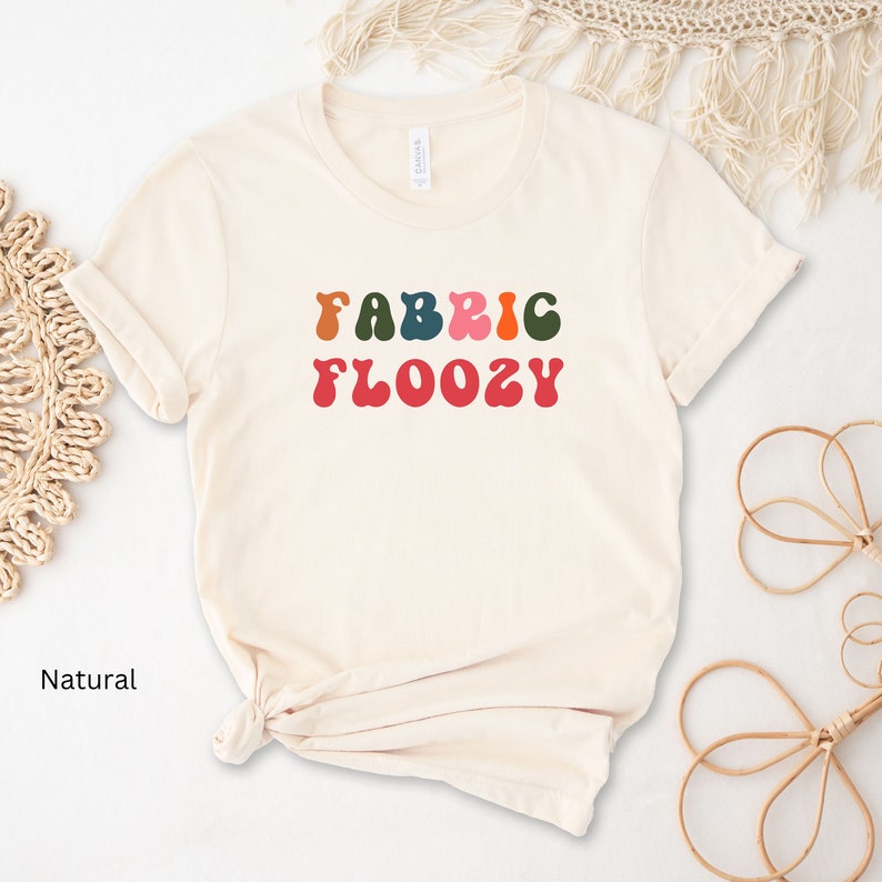 Pu&ograve; includere: Una maglietta color crema con la scritta "FABRIC FLOOZY" in lettere colorate in stile retr&ograve;. Il testo &egrave; nei toni dell'arancione, verde, blu, rosa e rosso. La maglietta &egrave; annodata in basso.