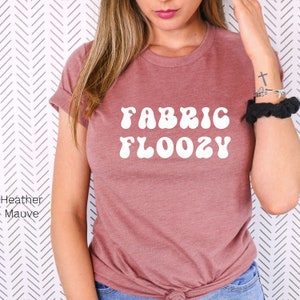 Pu&ograve; includere: T-shirt color malva con le parole "FABRIC FLOOZY" in bianco, con un font in stile retr&ograve;. La maglietta ha le maniche arrotolate ed &egrave; realizzata in un materiale morbido. La maglietta &egrave; di colore pieno e di stile casual.