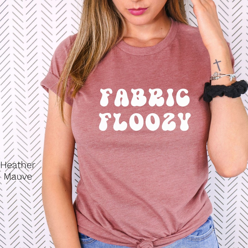 Pu&ograve; includere: T-shirt color malva con le parole "FABRIC FLOOZY" in bianco, con un font in stile retr&ograve;. La maglietta ha le maniche arrotolate ed &egrave; realizzata in un materiale morbido. La maglietta &egrave; di colore pieno e di stile casual.