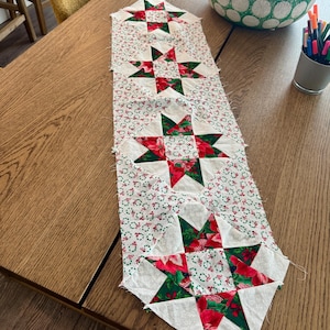 May include: A rectangular fabric table runner featuring a repeating pattern of four star-shaped quilt blocks. The blocks are constructed from white, red, and green fabric with floral and wreath prints. The runner has unfinished, raw edges.