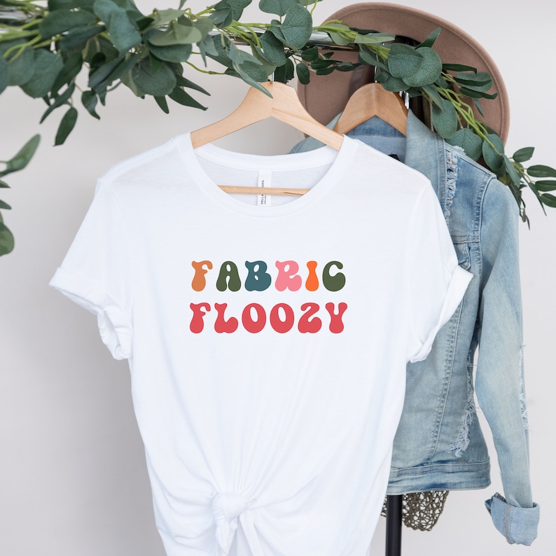 Pu&ograve; includere: T-shirt bianco con la scritta "FABRIC FLOOZY" in lettering colorato in stile retr&ograve;. La maglietta &egrave; appesa a una gruccia di legno, con una giacca di jeans e un cappello sullo sfondo. Il testo &egrave; arancione, rosa, verde e rosso.