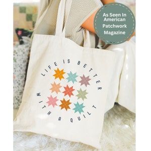 May include: A white canvas tote bag with the text "Life is better with a quilt" in blue lettering. The text is surrounded by a circle of colorful patchwork stars.