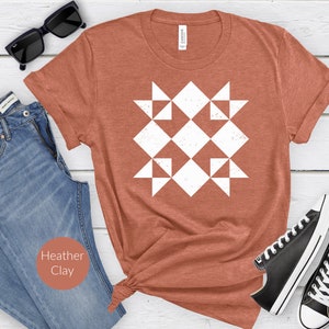 May include: A heather clay-colored t-shirt featuring a white geometric design. The design is a repeating pattern of squares and triangles, giving the appearance of a quilt block. The shirt is styled with blue jeans, black sunglasses, and black sneakers.