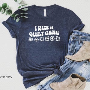 May include: Heather navy blue t-shirt with the text "I RUN A QUILT GANG" in white, along with decorative star and diamond shapes. The shirt is paired with blue jeans and tan ankle boots.