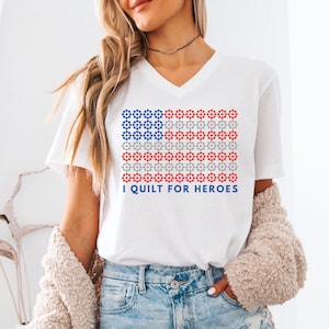 May include: White V-neck t-shirt with a red, white, and blue American flag design made of small stars. The text "I QUILT FOR HEROES" is printed below the flag.