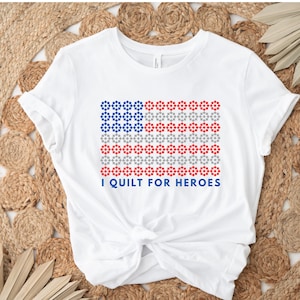 May include: White t-shirt with a red, white, and blue American flag design made of small stars. The text "I quilt for heroes" is printed below the flag.