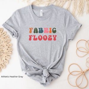 Pu&ograve; includere: T-shirt grigio atletico con la scritta "FABRIC FLOOZY" in lettere colorate in stile retr&ograve;. La maglietta &egrave; annodata in vita. La maglietta &egrave; realizzata in un materiale morbido.