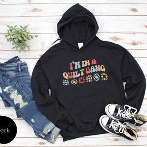 May include: Black hooded sweatshirt with the text "I'm in a Quilt Gang" in rainbow colors with a row of colorful quilt block designs below the text.