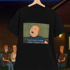 Puede incluir: Camiseta negra con una imagen de dibujos animados de Bobby Hill y la cita "THAT'S MY PURSE I DON'T KNOW YOU." La imagen es de la serie de televisión animada King of the Hill. La camiseta está colgada en una percha de madera.