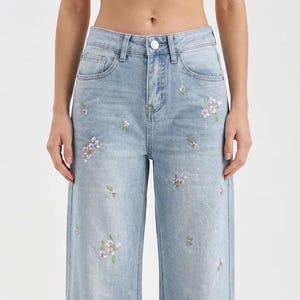 Op de afbeelding: Lichtblauwe jeans met wijde pijpen, versierd met geborduurde bloemmotieven in roze en wit. De jeans heeft een klassiek ontwerp met vijf zakken en een knoopsluiting. De lengte is ongeveer 100 cm.
