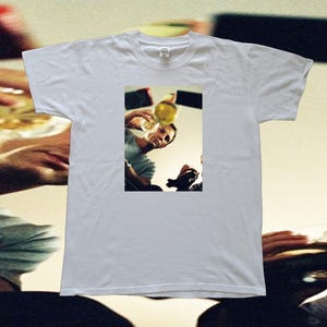 Peut inclure: T-shirt blanc avec une image rectangulaire d'une personne versant un liquide jaune d'une bouteille dans un verre. L'image est placée sur un fond neutre, suggérant un cadre décontracté ou social.