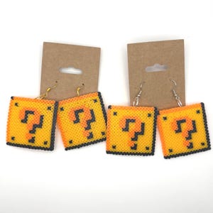 May include: Four pairs of square earrings with a yellow and black question mark design. The earrings are made of small, colorful beads. The earrings are attached to gold and silver hooks, and are displayed on brown cardboard.