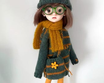 May include: A doll dressed in a knitted outfit, including a green hat with a yellow pom-pom, green-framed glasses, and a mustard-colored scarf. The doll wears a green and orange striped sweater dress, a matching purse with a flower, and boots.