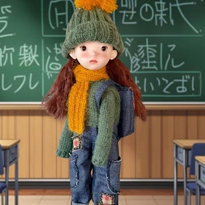 May include: A doll wearing a green knitted hat with an orange bobble, a green jumper, and denim dungarees. The doll has a yellow scarf and a small backpack. The background shows a classroom with a chalkboard and desks.