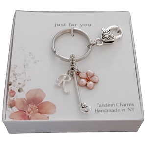May include: A silver keychain with a pink flower charm, a silver letter R charm, and a silver golf club charm. The keychain is attached to a silver heart-shaped clasp. The keychain is in a white box with the words "Just for you" and "Tandem Charms Handmade in NY" printed on it.