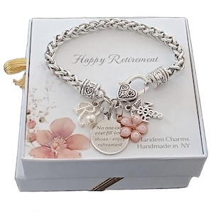 May include: A silver bracelet with charms, including a heart, a flower, a letter 'R', and a medical symbol. The bracelet is in a white box with the words 'Happy Retirement' and 'Tandem Charms Handmade in NY'. The charm with the flower has a pink center and white petals. The charm with the letter 'R' has a small pearl attached. The charm with the medical symbol has a red cross. The charm with the heart has a decorative design. The bracelet is in a white box with a pink floral design.