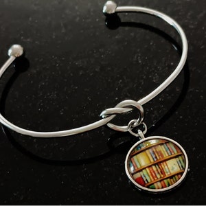 May include: A silver bangle bracelet with a knot design and a charm featuring a colorful image of books on a bookshelf.