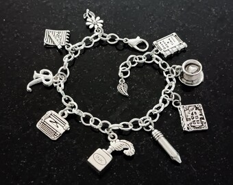 May include: A silver charm bracelet with various charms, including a typewriter, a book, a coffee cup, a pen, a flower, a leaf, a bottle of ink, and a notebook. The charms are attached to a silver chain.