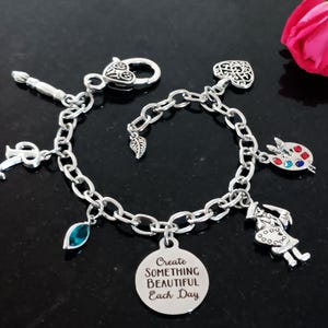 May include: A silver charm bracelet with a variety of charms, including a paint palette, a paintbrush, a leaf, a heart, a key, a teal blue gemstone, and a round charm with the text "Create Something Beautiful Each Day".