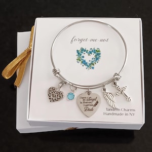 May include: A silver-tone wire bangle bracelet featuring four charms: a filigree heart, a light blue crystal, a heart-shaped pendant engraved with "I have an Angel in Heaven, I call him Dad," and a wing and cross charm, presented in a white gift box.