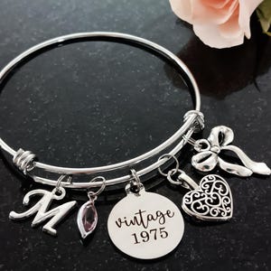 50th birthday 60mm stainless steel adjustable bangle bracelet with 'vintage 1975' laser engraved charm, filigree heart, delicate ribbon, and script initial and birthstone for personalization. Shown with flower upper right hand side.
