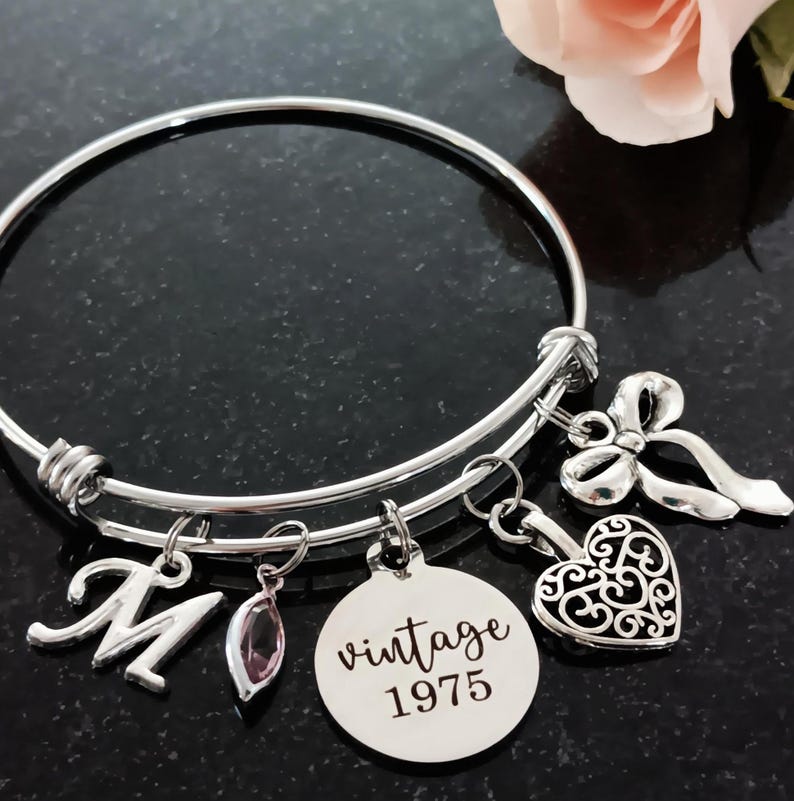 50th birthday 60mm stainless steel adjustable bangle bracelet with 'vintage 1975' laser engraved charm, filigree heart, delicate ribbon, and script initial and birthstone for personalization. Shown with flower upper right hand side.