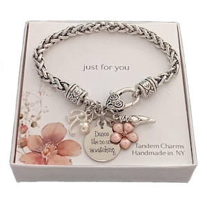 May include: A silver-toned braided chain bracelet featuring a heart-shaped clasp and multiple charms, including a round pendant engraved with "Dance like no one is watching," a letter R, a small pearl, a pink flower, and a ballet slipper charm.
