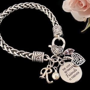 May include: A silver-toned chain bracelet featuring a lobster clasp, a heart-shaped charm, a small pink crystal, a letter R charm, a white pearl bead, and a round metal disc engraved with the text THE LOVE BETWEEN FRIENDS IS FOREVER.