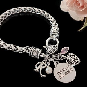 May include: Silver charm bracelet with a braided chain, a heart charm, a letter R charm, a pearl charm, and a round charm with the text "INTERIOR DESIGNER".