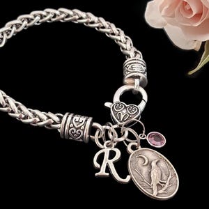 May include: A silver-toned wheat chain bracelet featuring a heart-shaped lobster clasp, two decorative metal beads, a letter R charm, an oval pendant depicting a bird on a branch under a crescent moon, and a small round pink crystal charm.