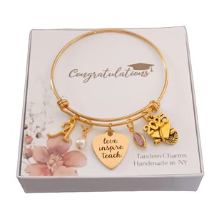 May include: Gold-toned bangle bracelet with multiple charms, including a heart pendant with the words "love, inspire, teach." Other charms include a graduation cap, the number "2025," and the letter "A." The bracelet is presented in a gift box.