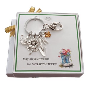 May include: A white gift box with a green border contains a silver keychain with charms. The charms include a watering can, a flower, a trowel, the letter "R", a bee, and a heart. The box has a green background with a floral design and the text "May all your weeds be WILDFLOWERS".