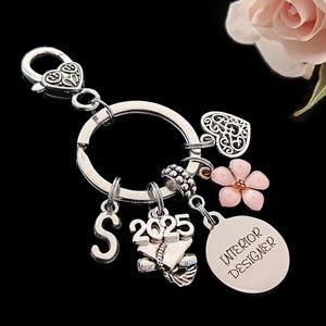 May include: Silver key chain with charms including a heart, flower, graduation cap, and a charm with the text "Interior Designer". The year 2025 is also included on the keychain.