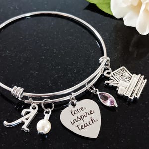 May include: Silver expandable bangle bracelet with charms. The charms include a heart with the text "love inspire teach", a book stack, a letter A, a pearl, and a purple crystal.