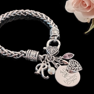 May include: Silver-toned charm bracelet with a braided chain design. The bracelet features multiple charms, including a heart-shaped lock, a letter "R", a pearl, a pink crystal, and a disc that reads "40 & Fabulous".