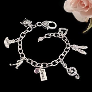 May include: A silver-toned charm bracelet featuring a lobster clasp and various dangling charms, including a butterfly, a heart, ballet slippers, a treble clef, the letter A, a pink crystal, and a rectangular tag with the word Dance engraved.