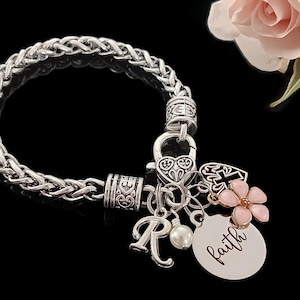 May include: A silver charm bracelet with a braided chain, multiple charms, and a pink flower charm. The charms include a heart, a cross, a letter "R", and a pearl. The bracelet also has a silver tag with the word "faith" engraved on it.
