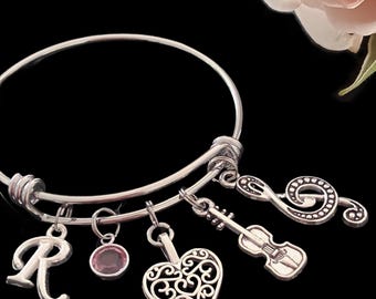May include: A silver-toned expandable bangle bracelet featuring five dangling charms: a letter R, a small round pink crystal, a filigree heart, a violin, and a treble clef. The bracelet is set against a black background with a pale pink rose.