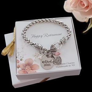 May include: A silver bracelet with a charm that says "retired 2025" and a pink heart charm. The bracelet is in a white box with the words "Happy Retirement" on it.